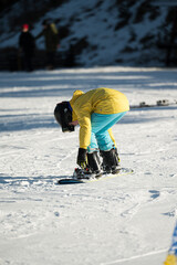 Young snowboarder adjusts bindings on a sunny winter day at a snowy mountain resort surrounded by...