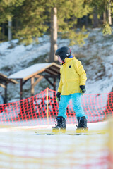 Brightly dressed young snowboarder learning to balance on a snowy slope surrounded by trees and ski...
