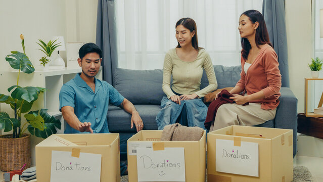Asian friends sorting and placing clothes into cardboard donation boxes at home. Concept of charity, social contribution, teamwork, and sustainable living.