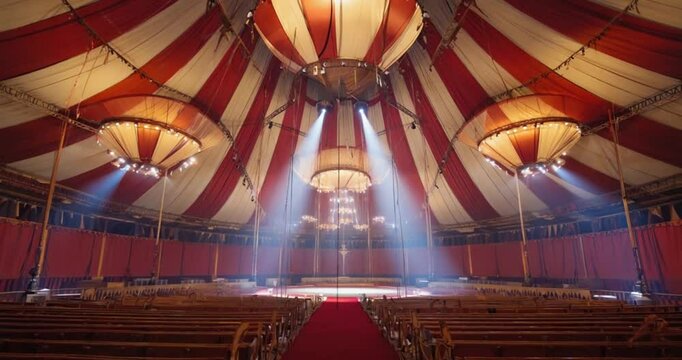 A grand indoor circus tent, with red and white striped canopies, ornate chandeliers, and rows of wooden seats