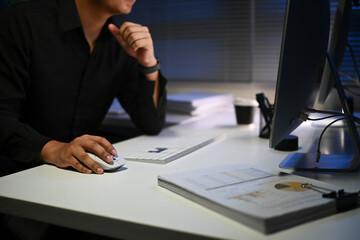 A man is sitting at a desk with a computer monitor in front of him at night. He is wearing a black...