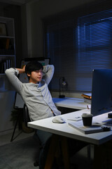 A young man is sitting at a desk with a computer monitor in front of him. He is wearing a white...