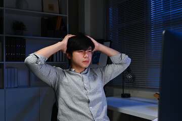 A man with his hands on his head in front of a computer monitor. He is wearing a gray shirt and he...