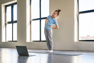 Fototapeta premium Young child practicing yoga at home during a sunny day with a laptop in view.