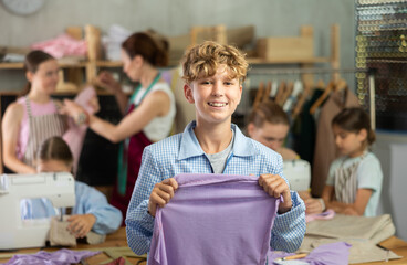 Cheerful preteen boy enjoying relaxed sewing workshop for children led by professional seamstress, proudly holding up stitched fabric while groupmates working in background