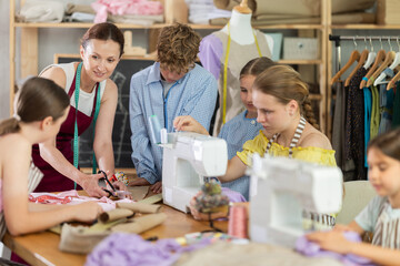 Professional friendly seamstress leading class of garment construction for interested teenage girls and boys in sewing workshop, demonstrating fabric cutting along pinned paper pattern..