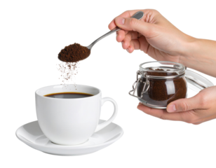 A hand sprinkles ground coffee from a spoon into a white cup filled with black coffee, with a glass jar of coffee grounds held in the other hand.
