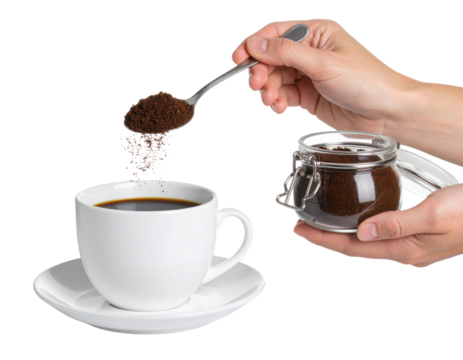 A hand sprinkles ground coffee from a spoon into a white cup filled with black coffee, with a glass jar of coffee grounds held in the other hand.