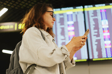 Young female tourist using smartphone checking flight schedule information board in airport