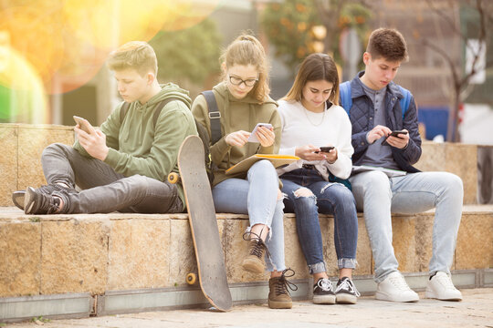 Group of boys and girls teenager addicted in their phones sitting on bench outdoors - Powered by Adobe