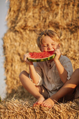 Close-up portrait of a cheerful boy biting a juicy red watermelon on a haystack in a sunny summer day. Natural light, childhood joy and healthy rural lifestyle concept