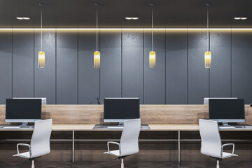 Modern dark interior office with wooden desk row, white chairs, and computers under pendant lights...