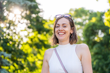 Cheerful young woman smiling brightly outdoors in sunlit park surrounded by vibrant green foliage...
