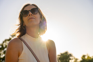Confident woman in sunglasses standing under evening sun in green park, sunlight flare highlights...