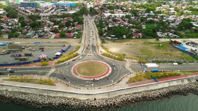 Sideways drone glide tracks a busy seaside roundabout in Davao City, seawall in foreground, tree-lined boulevard and colourful homes stretching inland, fairground rides to the left.