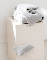 Stacked neatly white and gray towels on a minimalist white pedestal with a single towel on the floor in a bright, modern bathroom or spa setting