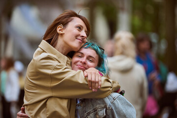Fototapeta premium Young adult females with brown and blue hair share warm embrace. Casual attire, smiles. Blurred urban background, soft lighting, relaxed atmosphere