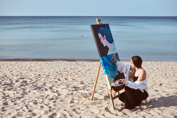 Young female artist in black dress paints portrait on canvas set against serene seaside. Scene...