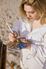 Young woman artist stands on sandy beach, engrossed in her painting. She holds colorful palette and...