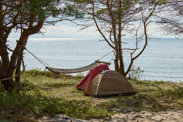 Two tents and hammock are set up beneath trees on tranquil beach. Scene embodies relaxation,...
