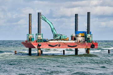 Offshore platform with green excavator on red base in ocean. Overcast sky and waves enhance...