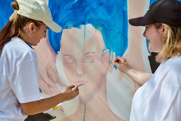 Young women working on large mural outdoors, bright colors. Focused expressions. Blue dominant,...