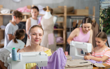 Smiling teenage girl holding piece of lavender fabric during sewing group class while classmates stitching on machines under guidance of female teacher in school workshop