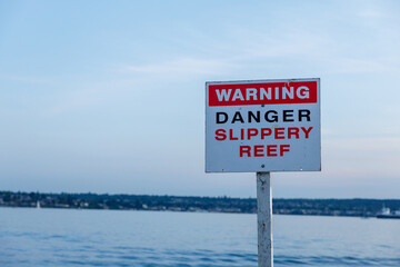 Warning sign indicating danger and slippery reef near a calm waterfront, with a ship visible in the...