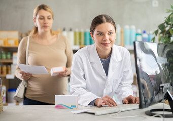 While working with client, girl pharmacist reads instructions for drug on computer, checks...