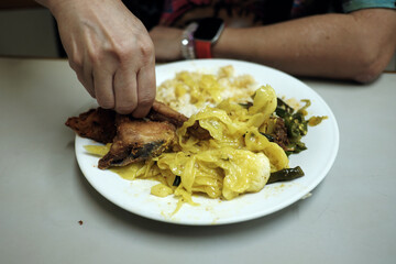 A hand reaches for a plate of Nasi Padang with gulai cabbage, grilled Padang-style chicken, and rice, typically enjoyed by hand.