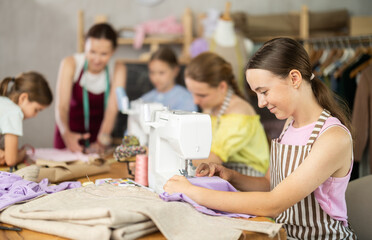 Teenage girl works on sewing machine while teacher and students learn how to cut fabric