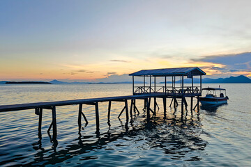 A peaceful wooden jetty at sunset on one of the islands near Semporna, Sabah, Malaysia. A small boat is moored gently on the calm sea as golden light bathes the horizon.