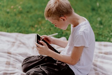 A boy with hearing aids plays on the grass while engaging with gadgets - drawing on a tablet and...