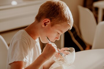 A boy with hearing aids enjoys a bowl of warm porridge in a sunlit kitchen, radiating pure...