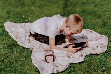 A boy with hearing aids plays on the grass while engaging with gadgets - drawing on a tablet and...