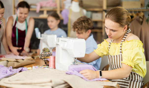 Teenage girl works on sewing machine while teacher and students learn how to cut fabric