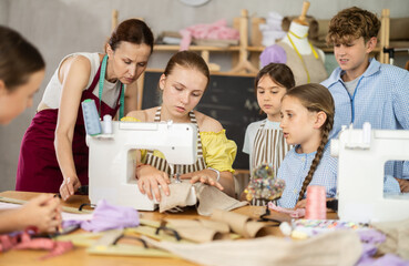Engaged schoolgirl about fifteen practicing stitching at machine under guidance of female...