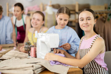 Teenage girl works on sewing machine while teacher and students learn how to cut fabric