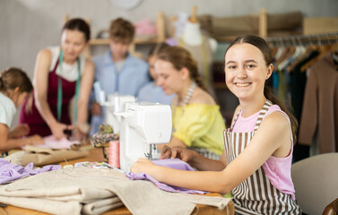 Smiling teen girl focusing on sewing on machine during hands-on lesson while classmates preparing...