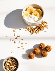 A bowl of sliced bananas with granola and yogurt, accompanied by scattered cereal oats and energy bites on a white background, highlighting healthy breakfast options