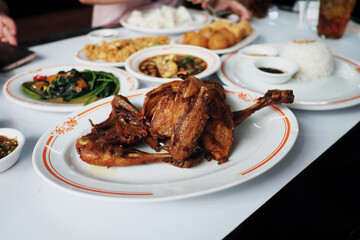 
A table laden with an Indonesian family meal, featuring a whole fried chicken, stir-fried greens, perkedel, and rice, set in a casual dining atmosphere.