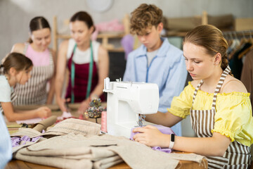 Teenage girl works on sewing machine while teacher and students learn how to cut fabric