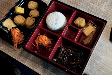 A Japanese-style bento box filled with rice, a dark main dish, omelet rolls, shredded carrots, and other sides, beside an extra tray of similar items.