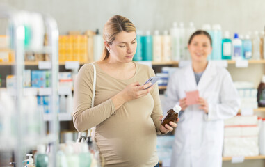 Modern pregnant woman using phone to scan QR code on bottle of syrup and check ingredients or...