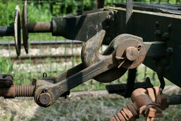 Rusty train hook coupler with large mechanical pivot, locking pin, metal springs, and corroded surface parts, photographed outdoors on railway tracks in daylight.