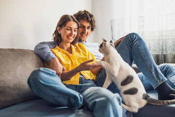 Portrait of young happy couple and their cat at home. They are holding paw of the cat.