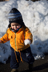 An adorable toddler dressed in a warm orange jacket and a striped hat stands in a snowy landscape,...