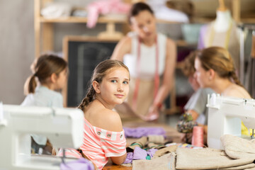 Smiling preteen girl sitting at table during school sewing workshop, while friendly, experienced...