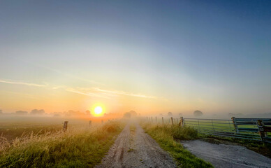 A serene and beautiful sunrise gently illuminates a foggy rural path, evoking feelings of deep...
