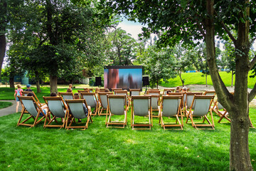 Summer outdoor cinema in a park. People sit on deck chairs watching a large screen, enjoying a film screening on a warm day.